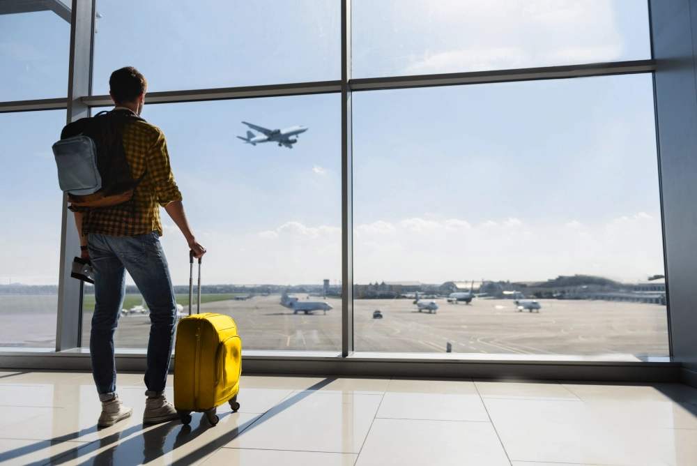 A man standing in front of a window in an airport, holding his suitcase and looking at a plane taking off.