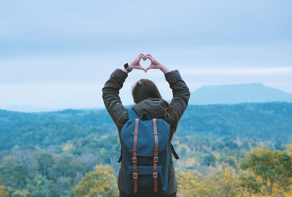 A person with a backpack forms a heart shape with their hands, symbolizing love and adventure in nature.